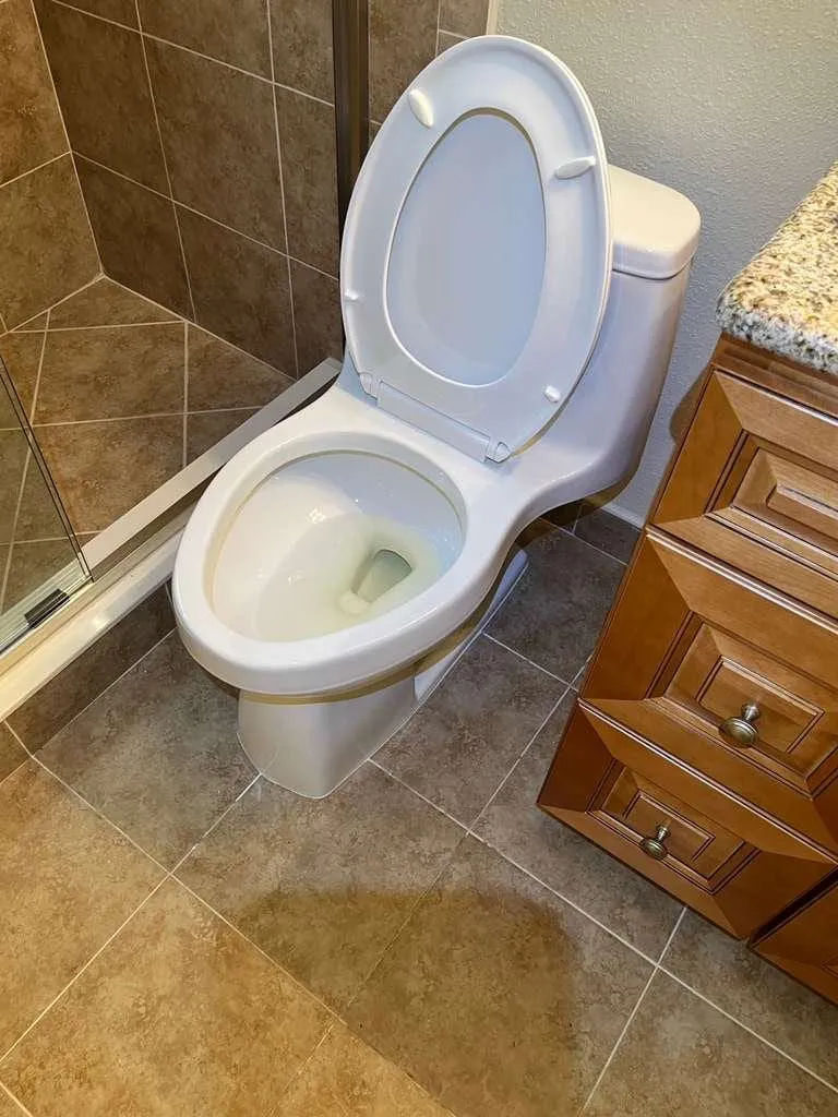 A toilet reset on beige tile beside a wood vanity, tank lid on and the work area wiped clean